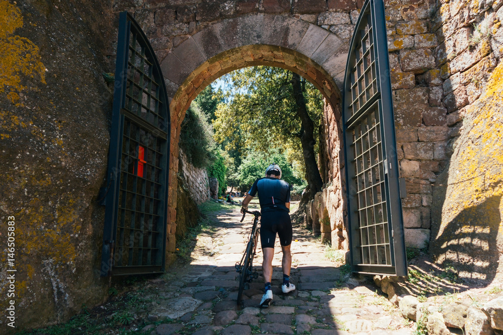 Strong male sprinter professional cyclist in midday sunlight, light from above, approaches open gate with iron and tan bricks in a high gothic village  in the spanish mountains.