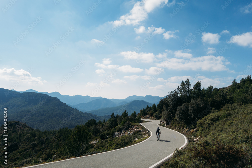 Fototapeta premium a fit young male cyclist riding on a windy spainiush mountain road through rocks and and very grren trees, soft white puffy clouds above and gently blue colored sky and hazed mountains.