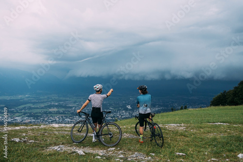 Two friends, teammates cyclists stopped, sitting on bikes, bicycles to view beautiful green lushious mountians with a giant rainstorm on a ride.