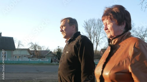 Loving elderly couple walking in the park at sunset. Nearby there is a road. A woman in a brown leather jacket. The man is dressed in a black sweater.