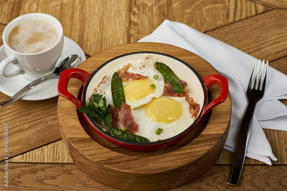 bacon and eggs with green peas in red pan and cup of americano coffee on wooden table in restaurant
