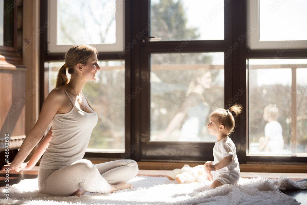 Young yogi mother at Easy Seat pose, cute baby daughter sitting near her, starting a new sporty day together, enjoying morning exercises at home, family fitness and fun time. Healthy lifestyle concept Young yogi mother at Easy Seat pose, cute baby daughter sitting near her, starting a new sporty day together, enjoying morning exercises at home, family fitness and fun time. Healthy lifestyle concept