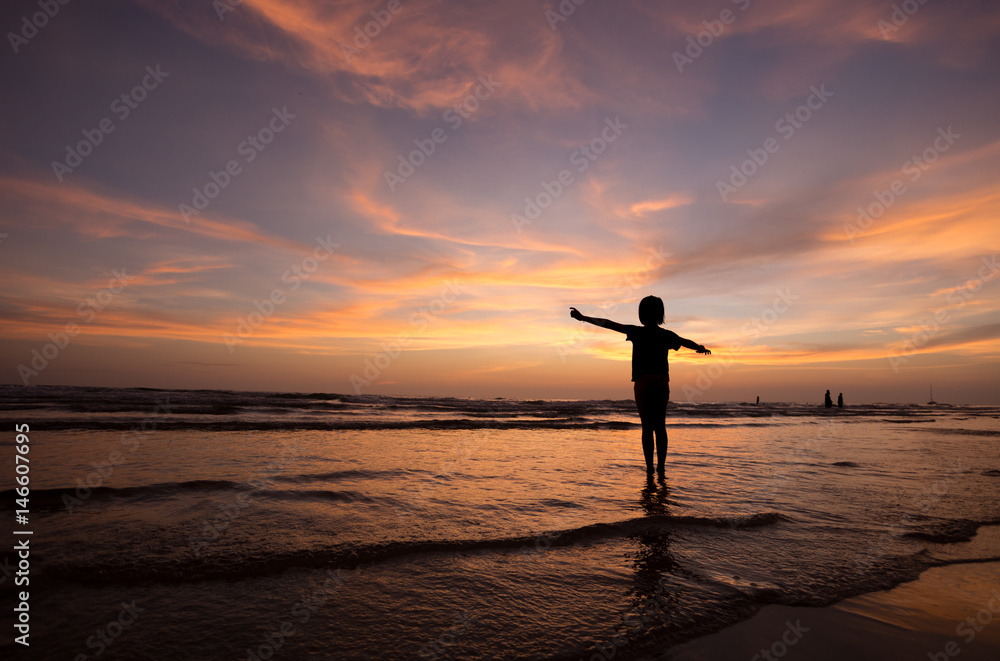 silhouette of happy girl  stand on sand of the sea and show hand in to sky with sunset. 