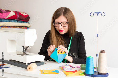 needlework and quilting of a young tailor - pretty woman tailor manually with needle stitching pieces of colorful fabrics at the workplace and thread, needles, a sewing machine and fabrics