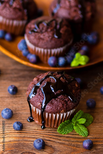 Chocolate muffins with chocolate syrup, blueberries and mint in a wooden background