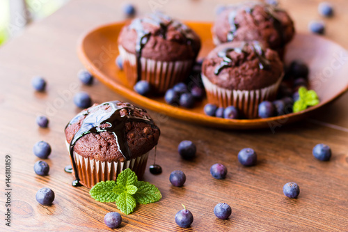 Chocolate muffins with chocolate syrup, blueberries and mint in a wooden background