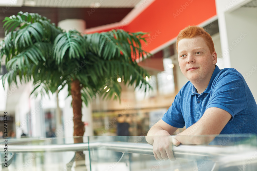 A photo of handsome stylish guy with trendy haircut and red hair dressed in casual blue T-shirt posing in a mall against big green palm tree. Thoughtful freckled boy with red hair having shopping.