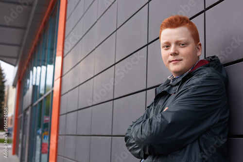 A horizontal portrait of redhead guy with freckles wearing black jacket posing over black wall of the shop. A handsome stylish guy with red hair standing crossed hands near the wall