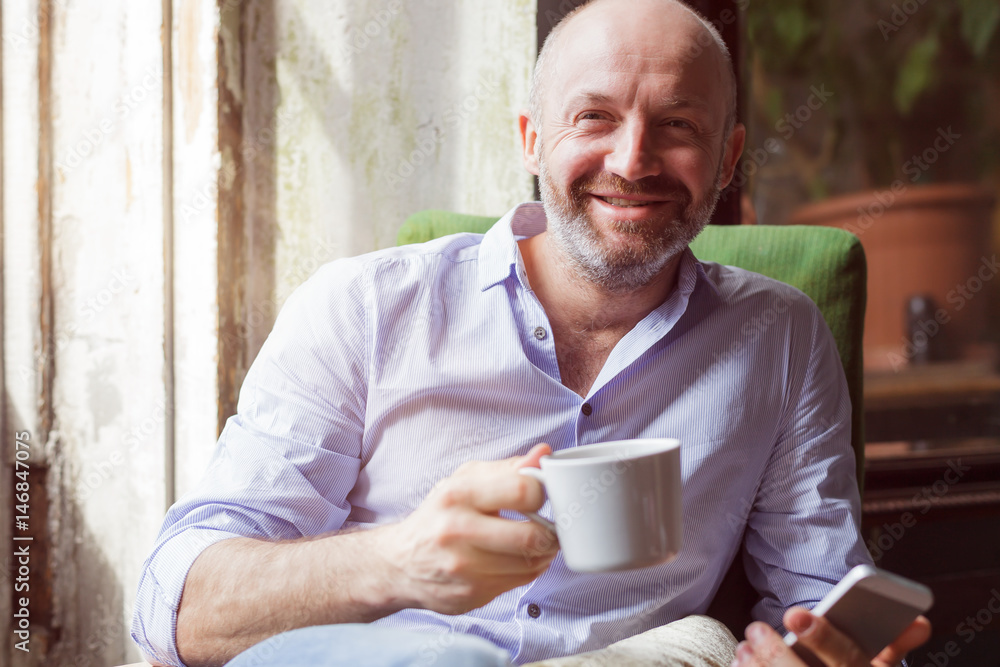 A man drinks coffee and checks the news in the phone, sitting in a chair by the window A man drinks coffee and checks the news in the phone, sitting in a chair by the window