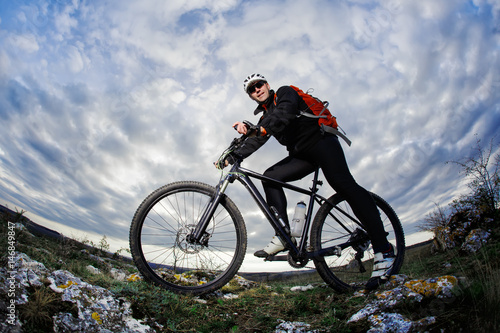 Photo below of the mountain cyclist in the black sportwear on the rocks against dramatic sky with clouds.