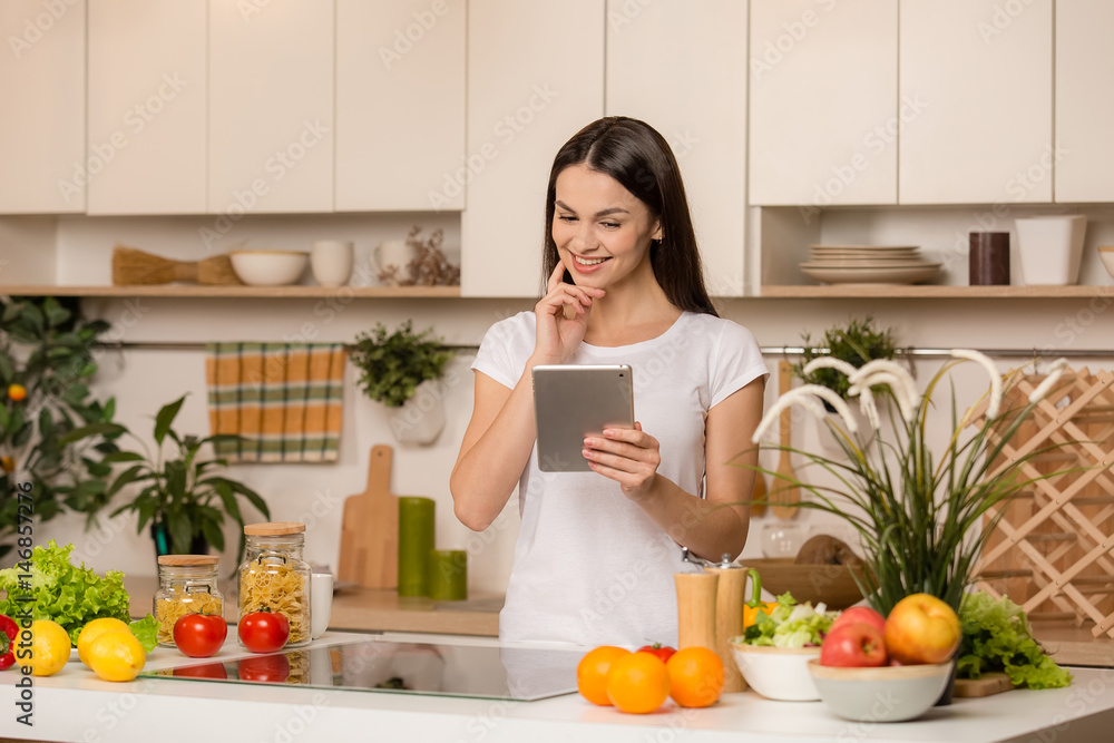 Young Woman in kitchen with tablet computer and looking recipes, smiling. Food blogger concept