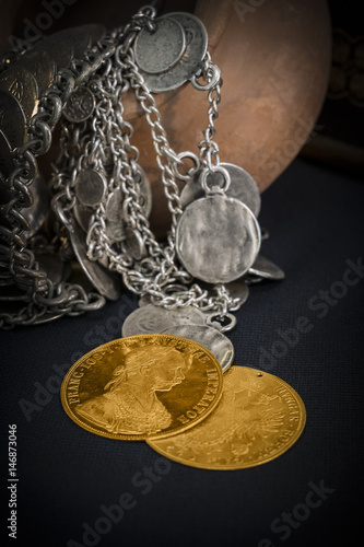 Still Life with two Austria-Hungary thalers, avers and revers of golden coin-ducats from 1915 with Kaiser Franz Joseph I, leaning on silver jewelery and dark environment photo