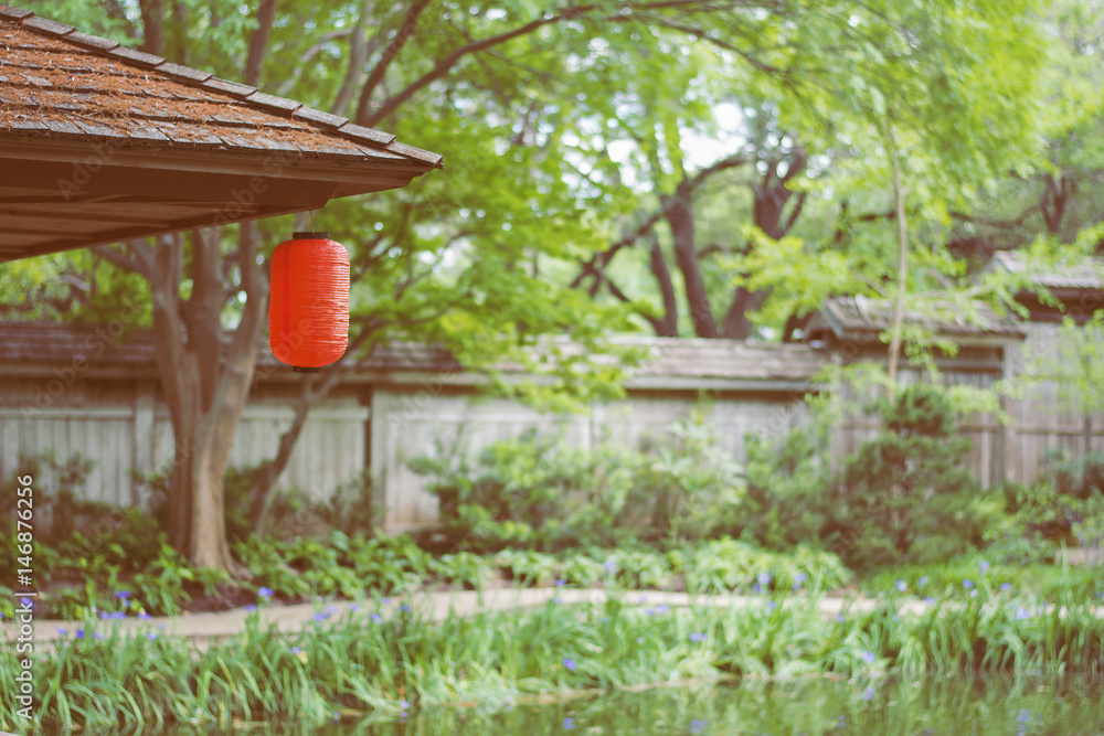 a red Japanese lantern hanging on the wooded roof  with green trees background, filtered tones