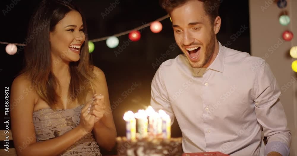 Young man blowing out the candles on his birthday cake watched by an excited laughing young woman with a string of party light decorations Young man blowing out the candles on his birthday cake watched by an excited laughing young woman with a string of party light decorations