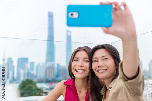 Chinese mother and daughter taking selfie together. Happy multiracial family on travel holiday in Shanghai, China. Mom holding mobile phone on outdoor cafe terrace with view of the Bund and Pudong. photo