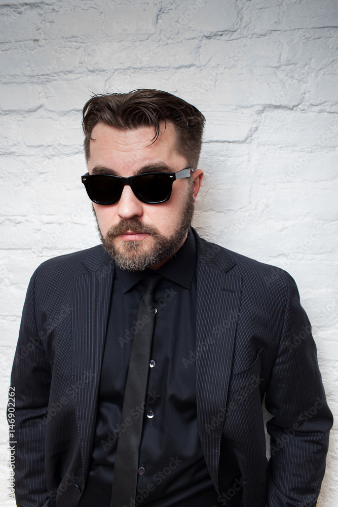 Obraz premium Portrait of a bearded serious man in sunglasses and a business black suit against a white brick wall