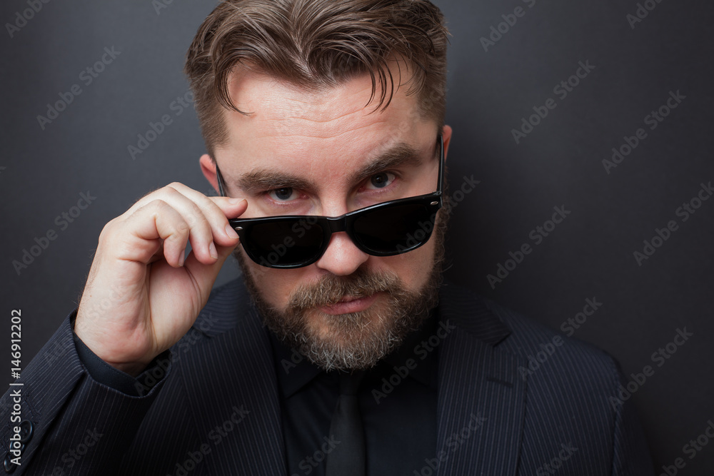A brutal man with a beard and a stylish hairstyle in a black suit looks appallingly at the camera, dropping his sunglasses A brutal man with a beard and a stylish hairstyle in a black suit looks appallingly at the camera, dropping his sunglasses