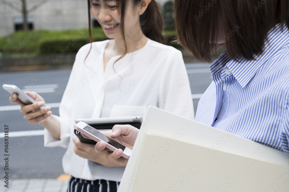 Two female office workers are talking outside while looking at the smartphone Two female office workers are talking outside while looking at the smartphone