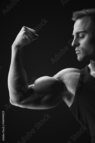 Black and white photo of young confindent muscular bodybuilder standing on black background and posing biceps muscle Classic bodybuilding