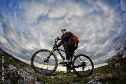 Portrait of the young cyclist standing with bike on the rocks against dramatic sky with clouds.
