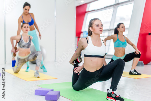 Group of women doing kneeling quads stretch exercise while fitness instructor helping in the background in studio