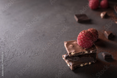 Broken chocolate pieces and raspberries on metal plate on brown background with copy space. Dark photo.