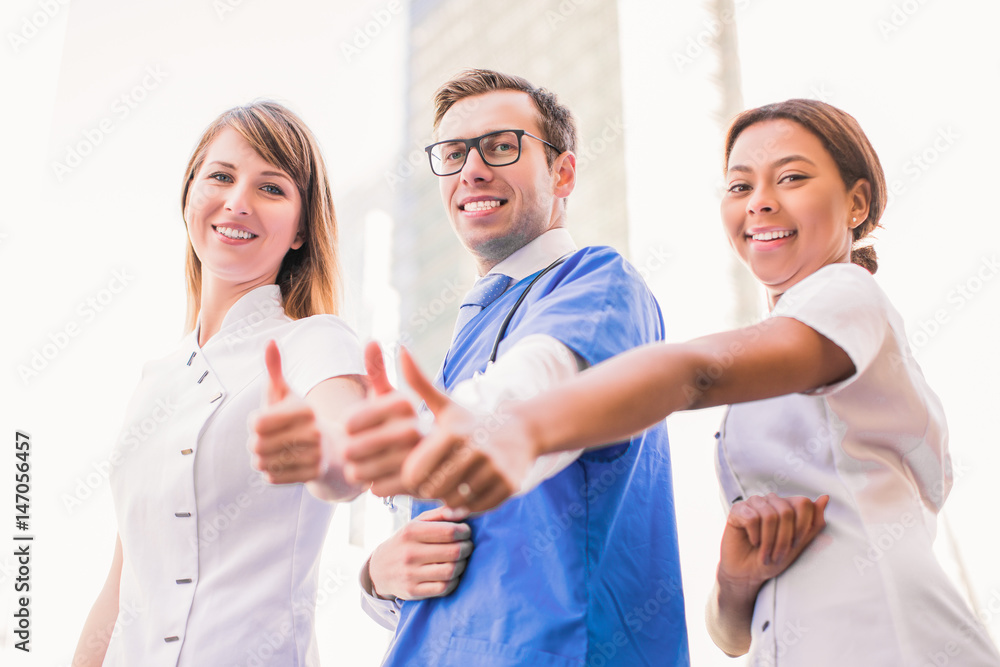 Three smiling happy doctors stand and show thumbs up outdoor Three smiling happy doctors stand and show thumbs up outdoor