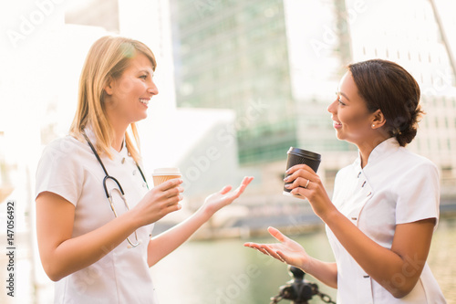Two smiling nurses stand with cups of coffee and tell with each other during the coffee break, outdoors