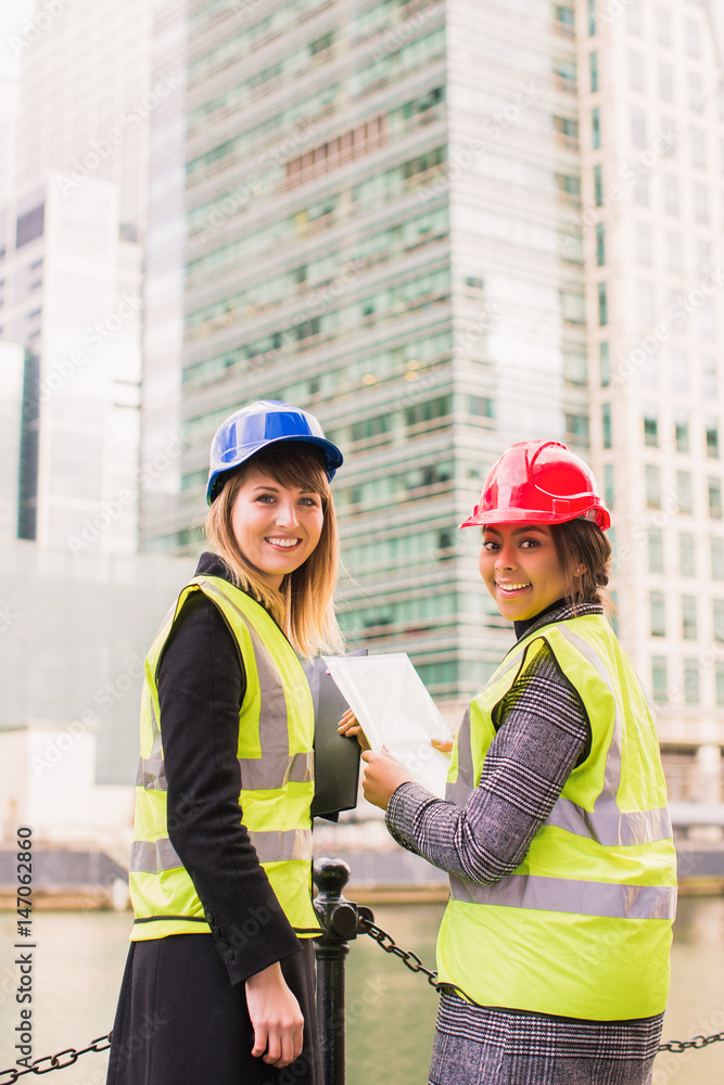 Portrait of two young, smiling women architects who stand together and keep some documents in hands, outdoors Portrait of two young, smiling women architects who stand together and keep some documents in hands, outdoors