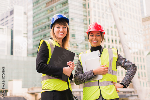 Portrait of two young, women architects who stand together, smile and keep the folder with some documents and the tablet in hands, outdoors