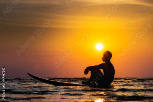 Silhouette of surf man sit on a surfboard. Surfing at sunset beach. Outdoor water sport adventure lifestyle.Summer activity. Handsome Asia male model in his 20s.