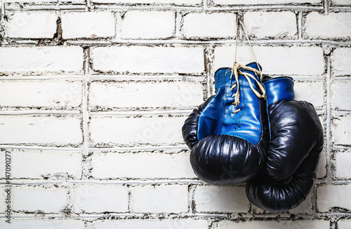 Pair of old blue and black boxing gloves hanging on white brick wall. © Veresovich