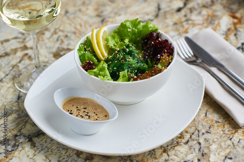 Seeweed salad in white ceramic bowl served with lemon slices, sauce, glass of white wine, fork and knife on marble table