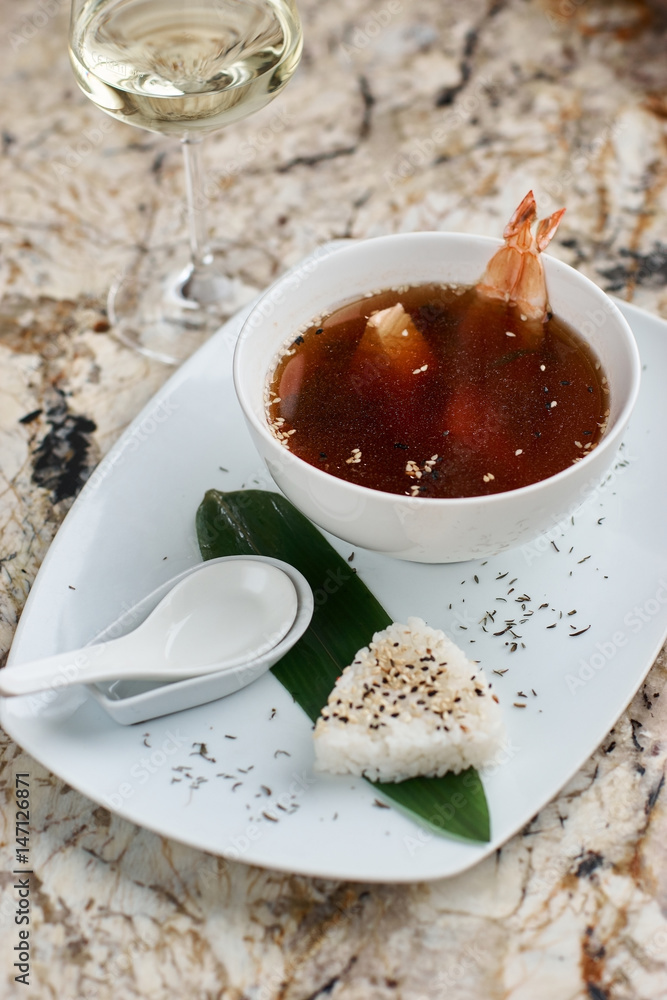 Japanese cuisine. Suimono or Matsutake soup served in white ceramic plate with spoon, rice and white wine on marble background. Japanese cuisine. Suimono or Matsutake soup served in white ceramic plate with spoon, rice and white wine on marble background.