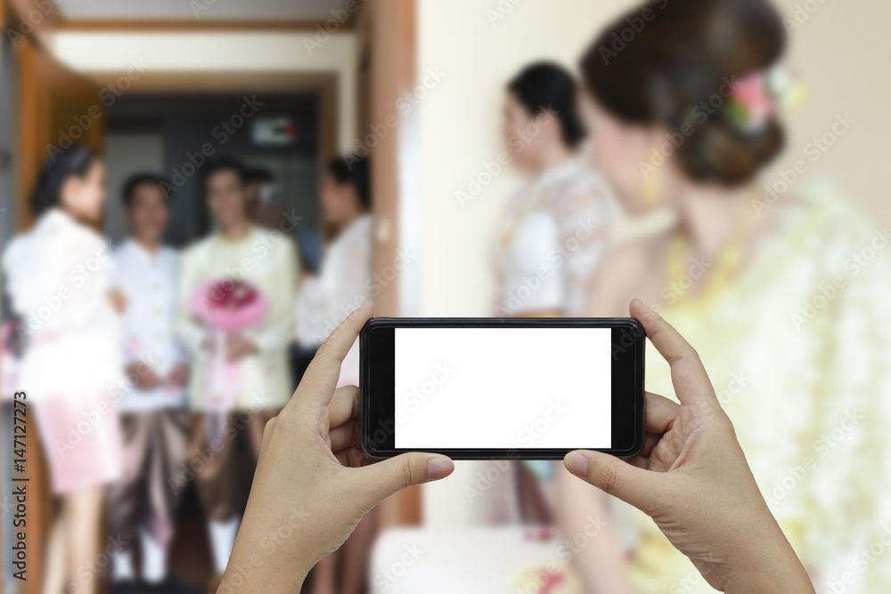 Hand holding smartphone with white blank screen and blurred wedding scene in the room background.