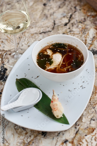Fototapeta Naklejka Na Ścianę i Meble -  Japanese cuisine. Noodle soup udon in white ceramic bowl served with spoon, glass of white wine and king prawn on marble table