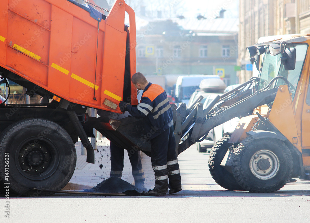 Fototapeta premium The dump truck pours asphalt crumb into the excavator bucket during extreme road repairs while the cars are moving.