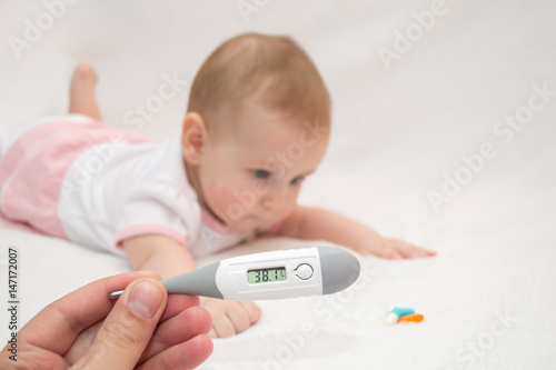 A hand holds a digital medical thermometer with a high temperature on a white isolated background with a child lying next to the medication tablets photo