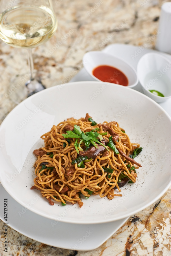 Flat egg noodles with duck meat, chicken, shiitake mushrooms served on white plate with white garlic and red spicy sauce Flat egg noodles with duck meat, chicken, shiitake mushrooms served on white plate with white garlic and red spicy sauce
