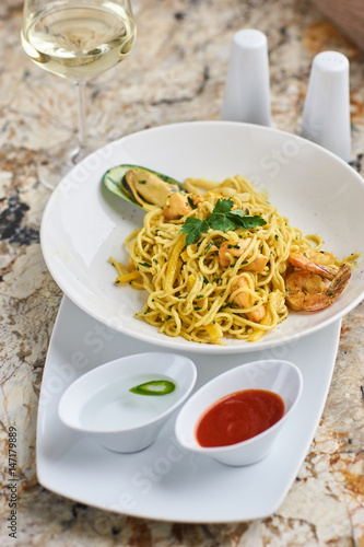 Flat egg noodles with seafood served in big white ceramic round plate with glass of white wine on marble table