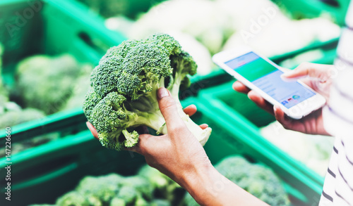 Young woman shopping healthy food in supermarket blur background. Female hands buy products broccoli using smartphone in store. Hipster at grocery using smartphone. Person comparing price of produce