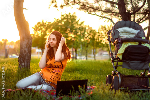 beautiful a red hair woman using computer and talking on smart phone sitting on grass in outdoor park on the sunset with sleeping baby in carrige photo