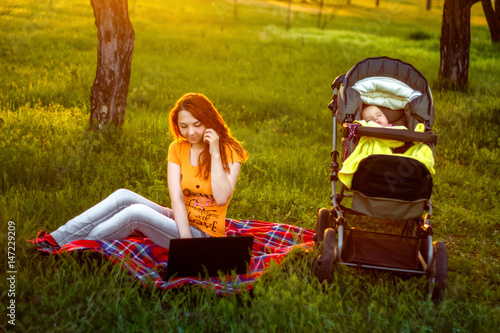 beautiful a red hair woman using computer and talking on smart phone sitting on grass in outdoor park on the sunset with sleeping baby in carrige photo