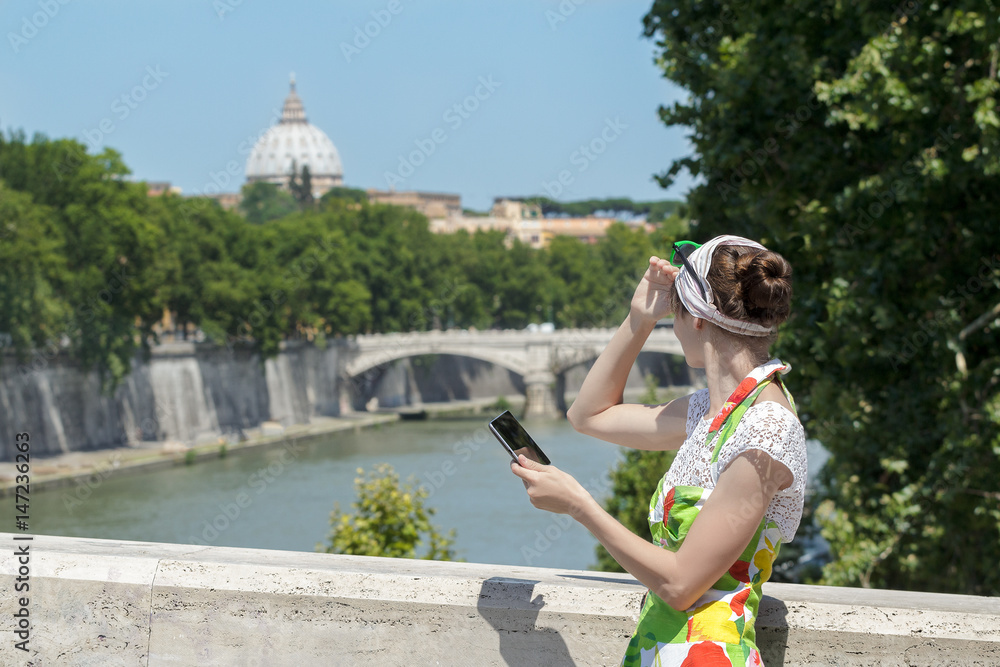 Tourist woman in flower sundress is covering eyes from the sun and holding tablet at Rome bridges and masterpiece dome background