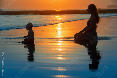 Family swimming fun on black sand beach with sea surf. Happy mother  baby son sit and look at ocean on sunset sky with sun background. Child with parents activity  healthy lifestyle on summer holiday.