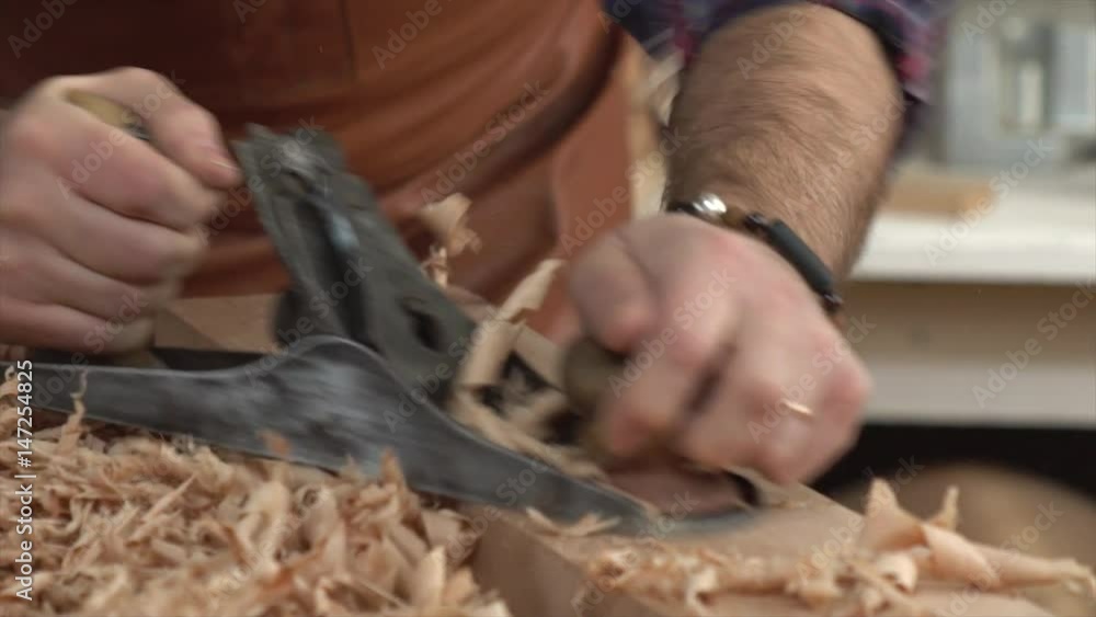 Cabinet-Maker Planes Solid Wood in his Workshop/Cabinet maker planes the beech plank with a plane. He works in a small family joiner’s shop Cabinet-Maker Planes Solid Wood in his Workshop/Cabinet maker planes the beech plank with a plane. He works in a small family joiner’s shop