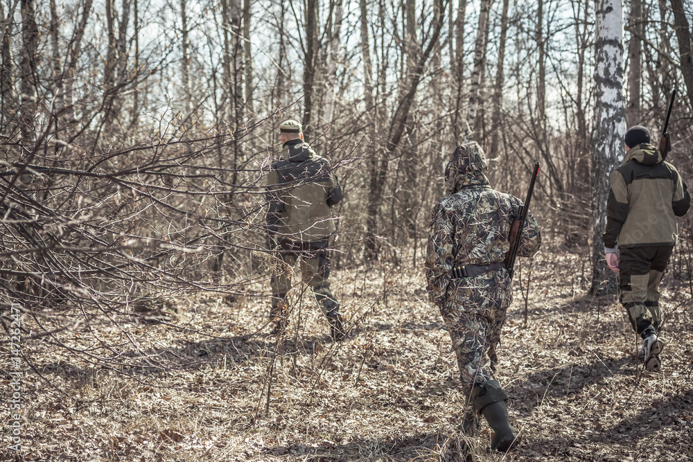 Hunting scene with group of hunters in camouflage walking in spring forest with dry leaves during hunting season