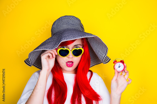 Attractive surprised red-haired young woman in sunglasses and hat on yellow background holding clock