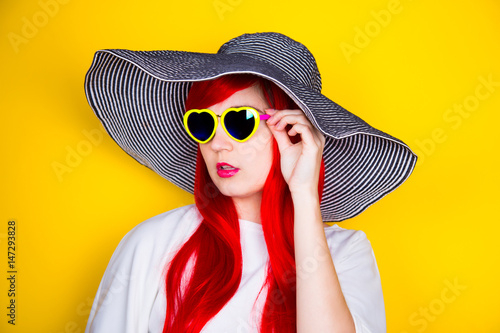 Attractive red-haired young woman in sunglasses and hat on yellow background