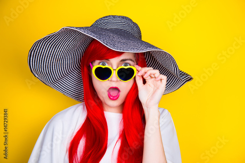 Attractive surprised red-haired young woman in sunglasses and hat on yellow background